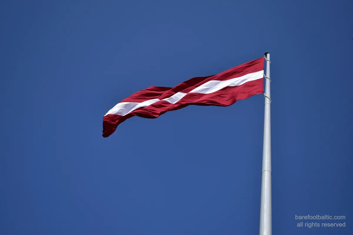 The Latvian flag — carmine red with a white band — flying against a blue sky.