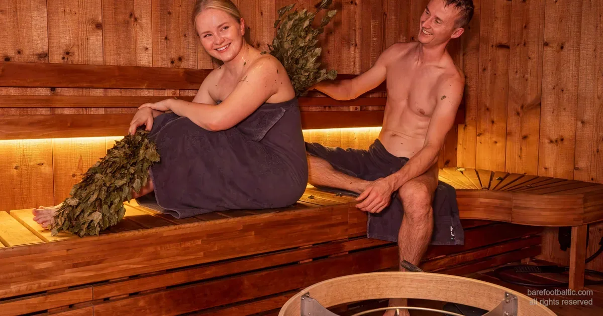 Wood-clad Baltic sauna interior — stone-filled kiuas heater and a birch vasta on the bench.